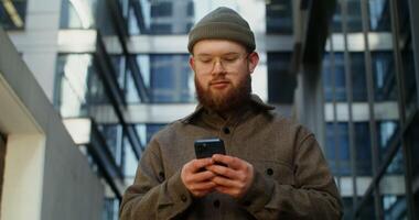 A young man in a knitted hat with a voluminous red beard is typing on a mobile phone while walking around the business center of the city, close-up. in 4k, red komodo video