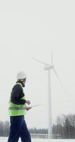 A young African-American man in a work uniform with a helmet on his head is diagnosing a wind turbine using a digital tablet, vertical . in 4k, red komodo video