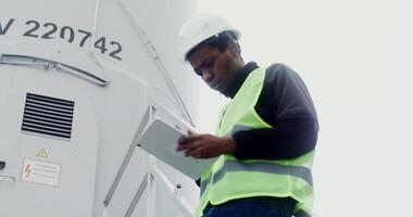A power plant worker in work gear uses a tablet with a serious look while standing on the steps near the entrance to an industrial wind generator. in 4k, red komodo video