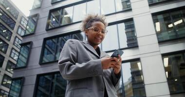 African-American woman dressed in business style with a pierced nose and wearing glasses smiles while using mobile phone while standing near office building, viewed from below. in 4k, red komodo video