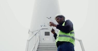 An African-American power plant worker in a green vest and hard hat uses a tablet with a serious look while standing near industrial wind turbine, conducting an activity check. in 4k, red komodo video