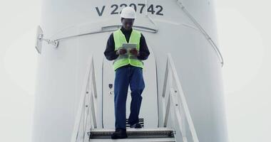 A young African-American man in work gear uses a tablet with a serious look as he walks down the steps of a wind turbine during an inspection. in 4k, red komodo video