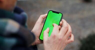 A warmly dressed man uses a phone with a green screen, swiping his finger across the screen, close-up, no face, view from behind the man. Copy space, mockup. in 4k, red komodo video