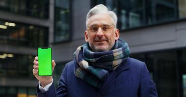 A solid warmly dressed middle-aged businessman smiling looking at camera holding the phone in his hands with the screen facing forward. Green screen, copy space, chroma key. in 4k, red komodo video