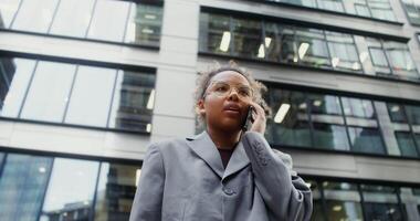 A young African-American woman dressed in a business suit with a serious look talking on a mobile phone while standing near an office building, bottom view, close-up. in 4k, red komodo video