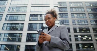 Young African-American woman dressed in business style smiling while using a mobile phone while standing near an office building, bottom view, dynamic shot. in 4k, red komodo video