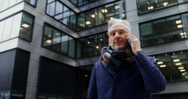 Mature man with a scarf around his neck, looking at camera and smiling while talking on a mobile phone, standing near an office building. from below, circular shooting. in 4k, red komodo video