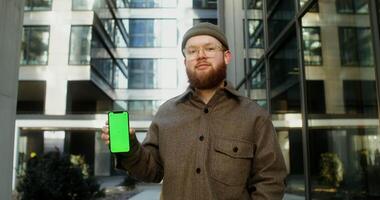 Young man with a beard dressed in casual clothes and a knitted hat smiling while looking at the camera, holding a mobile phone with a green screen, copy space, mockup. in 4k, red komodo video