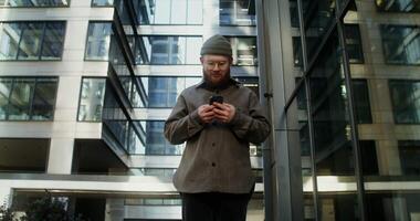 A young man with a beard, dressed in casual clothes and a knitted hat, smiles while typing on a mobile phone, standing in the courtyard of a modern glass building. in 4k, red komodo video