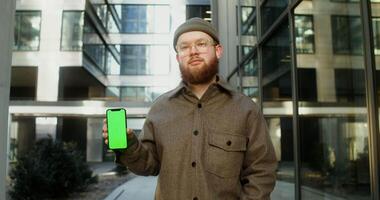 Mobile phone face forward in the hands of a young man dressed in casual clothes, standing in the business center of the city. Copy space, green screen, chroma key, mockup. in 4k, red komodo video