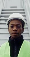 A young African-American man in a white hard hat and green vest turns his head to look at the camera and smiles, standing near the entrance to the wind turbine, vertical . in 4k, red komodo video