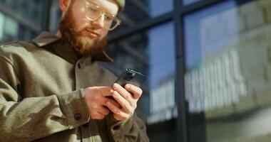 A young man in a knitted hat with a voluminous red beard is typing text on a mobile phone, standing near a glass building on a sunny day, close-up. in 4k, red komodo video