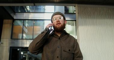 A young man with a beard, dressed in casual clothes and a knitted hat, is talking on a mobile phone while walking in the courtyard of a modern glass building, close-up. in 4k, red komodo video