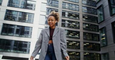 A young African-American woman wearing headphones, dressed in a business jacket over a crop top, dances rhythmically with enjoyment near a modern office building. in 4k, red komodo video