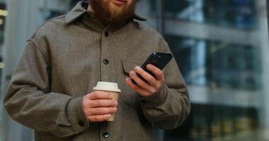A young bearded man in glasses is typing on a mobile while standing near office building with a disposable cup in his hands. Close-up, motion from hands to face. in 4k, red komodo video