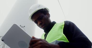 An African-American power plant worker in a green vest and hard hat uses a tablet with a serious look while standing near an industrial wind generator, close-up. in 4k, red komodo video