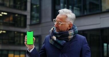 A handsome, warmly dressed, middle-aged businessman smiles at the camera while holding a green screen phone as he stands outside an office building. Copy space. in 4k, red komodo video