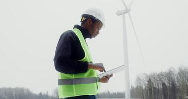 A young African-American man in a work uniform in a helmet on his head is engaged in the diagnosis of a wind turbine using a digital tablet, close-up, dynamic . in 4k, red komodo video