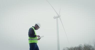 A young African-American man in a work uniform with a helmet on his head is diagnosing a wind turbine using a digital tablet while standing on the street near a wind turbine. in 4k, red komodo video