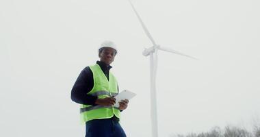 African-American man in work uniform with hard hat on his head smiles while looking at the camera while standing near a wind turbine on a winter day with a tablet in his hands. in 4k, red komodo video