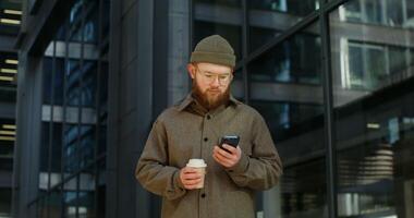 A young bearded stylishly dressed man smiling while using a mobile phone while standing near an office building with large darkened windows. in 4k, red komodo video