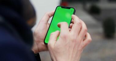 Close-up of a mobile phone with a green screen in the hands of an unknown man scrolling the screen. Mockup, copy space, view from behind a man. in 4k, red komodo video