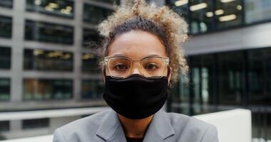 Young businesswoman of African American appearance in a medical mask on her face in glasses looks at the camera while standing near the modern building, close-up. in 4k, red komodo video