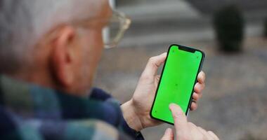 An adult gray-haired warmly dressed man uses a phone, swiping his finger across the green screen, close-up, top view from behind the man's shoulder. Chroma key, mockup. in 4k, red komodo video