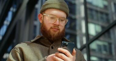 A young man in a knitted hat with a voluminous red beard is typing on a smartphone, standing near a glass building, close-up. in 4k, red komodo video