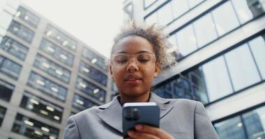 Young African American woman with nose piercing and glasses smiling while using mobile phone while standing near office building, close-up view from below. in 4k, red komodo video