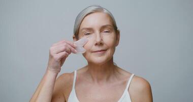 A professional elderly Caucasian model without makeup, with gray hair pulled back in a low ponytail, doing a facial massage using a Gua Sha stone while looking at the camera. 4K , red komodo video