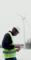 A young African-American man in a work uniform with a hard hat on his head is diagnosing a wind turbine using a digital tablet on a winter day, close-up, vertical . in 4k, red komodo video