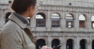 Young beautiful woman of European appearance smiling while typing a message on a mobile phone while standing against the backdrop of the Coliseum in Rome. in 4k, red komodo. video