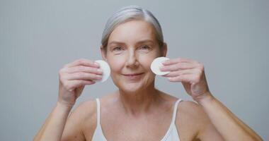 A fair-skinned older model with gathered gray hair wipes her make-up with cotton pads and smiling while looking straight into the camera, against a solid color studio background. 4K , red komodo video