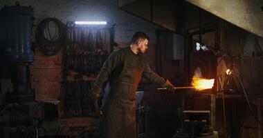 A blacksmith in protective gear and goggles shapes a red-hot metal blank with a hammer. Work in a modern forge. 4k , red komodo video