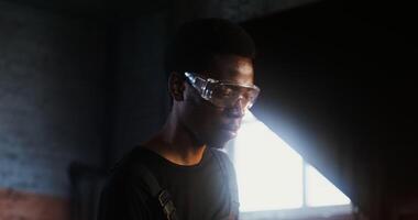 A young African-American man wearing clear goggles and a leather apron and gloves puts a metal bar into a red-hot furnace with tongs while working in a forge. in 4k, red komodo video
