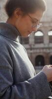 Young warmly dressed woman typing a message on her mobile phone while standing against the backdrop of the Coliseum in Rome. in 4k, red komodo, vertical video