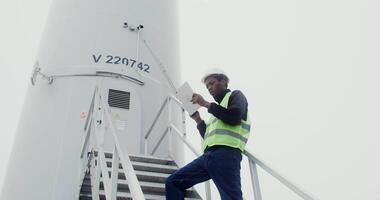 An African-American power plant worker in a green vest and hard hat uses a tablet with a serious look while standing on steps near the entrance to an industrial wind turbine. in 4k, red komodo video