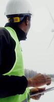 African-American man in a work uniform with a hard hat on his head uses a tablet while standing near a wind turbine and looking at it, winter day, close-up, vertical . in 4k, red komodo video