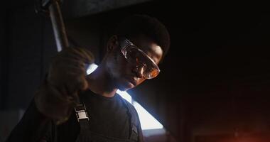 A young African-American man in goggles and gear uses a hammer to work hot metal while working in a forge. in 4k, red komodo video