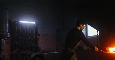 A young man in goggles and a leather apron takes a split metal blank out of the furnace with tongs and starts shaping the product with a hammer, working in a modern workshop. in 4k, red komodo video
