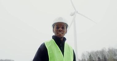 A young African-American man in a work uniform with a hard hat on his head smiles while looking at the camera, standing near a wind turbine. in 4k, red komodo video