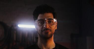 Young handsome bearded man in goggles looks directly at the camera while standing in a modern blacksmith workshop, close-up. in 4k, red komodo video