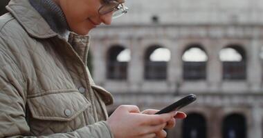 Young beautiful warmly dressed woman smiling while typing a message on her mobile phone while standing against the backdrop of the Coliseum in Rome. in 4k, red komodo. video