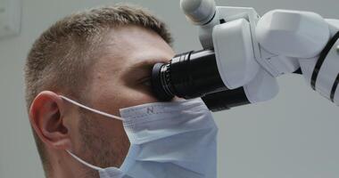 A young male doctor or laboratory assistant in a medical mask looks through a microscope while working in a laboratory, close-up. in 4k, red komodo. video