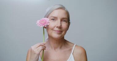 An aged caucasian model, without makeup, with gathered gray hair, posing with a gerbera flower, stroking her face and looking at camera, against a monochrome studio background. 4K , red komodo video