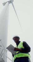 A young African-American man in a green vest and hard hat uses a tablet while standing near an industrial wind turbine, vertical , bottom view. in 4k, red komodo video