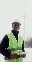 A young African-American man in work uniform with hard hat on his head smiles while looking at camera while standing near a wind turbine on a winter snowy day, vertical view. in 4k, red komodo video