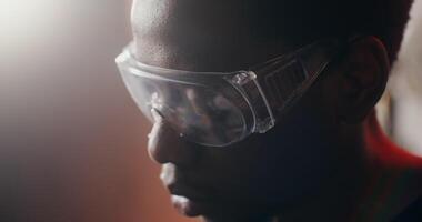 A young African-American man in protective transparent glasses looks down with a focused look while working in a workshop, close-up. The man's face in profile. in 4k, red komodo video
