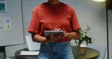 Young african american woman uses a digital tablet while leaning against a table in a meeting room in a stylish office, then looking up from the screen and smiling looking straight into the camera video